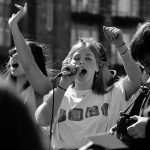 Una foto in bianco e nero con in primo piano una giovane che parla con un microfono durante una manifestazione