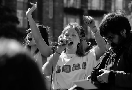 Una foto in bianco e nero con in primo piano una giovane che parla con un microfono durante una manifestazione